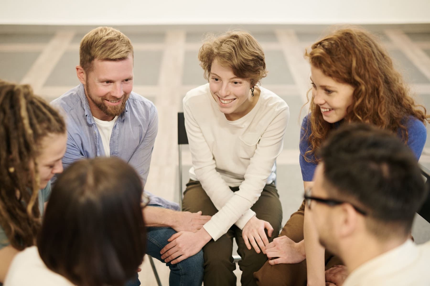Six adults sit in a circle on chairs, smiling and talking in a bright room