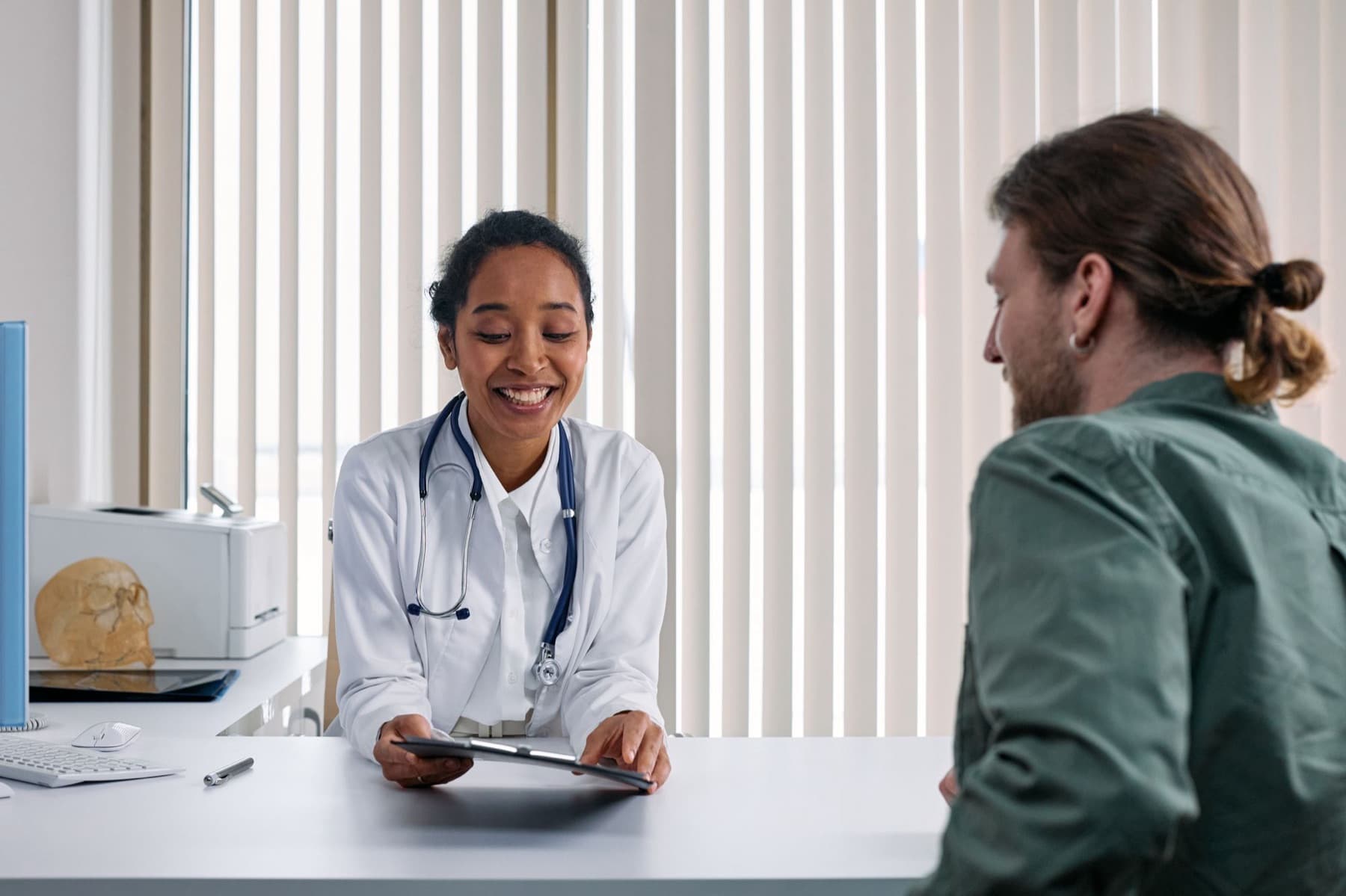 A smiling doctor with a stethoscope shows a patient notes on a tablet across a desk in a bright consulting room