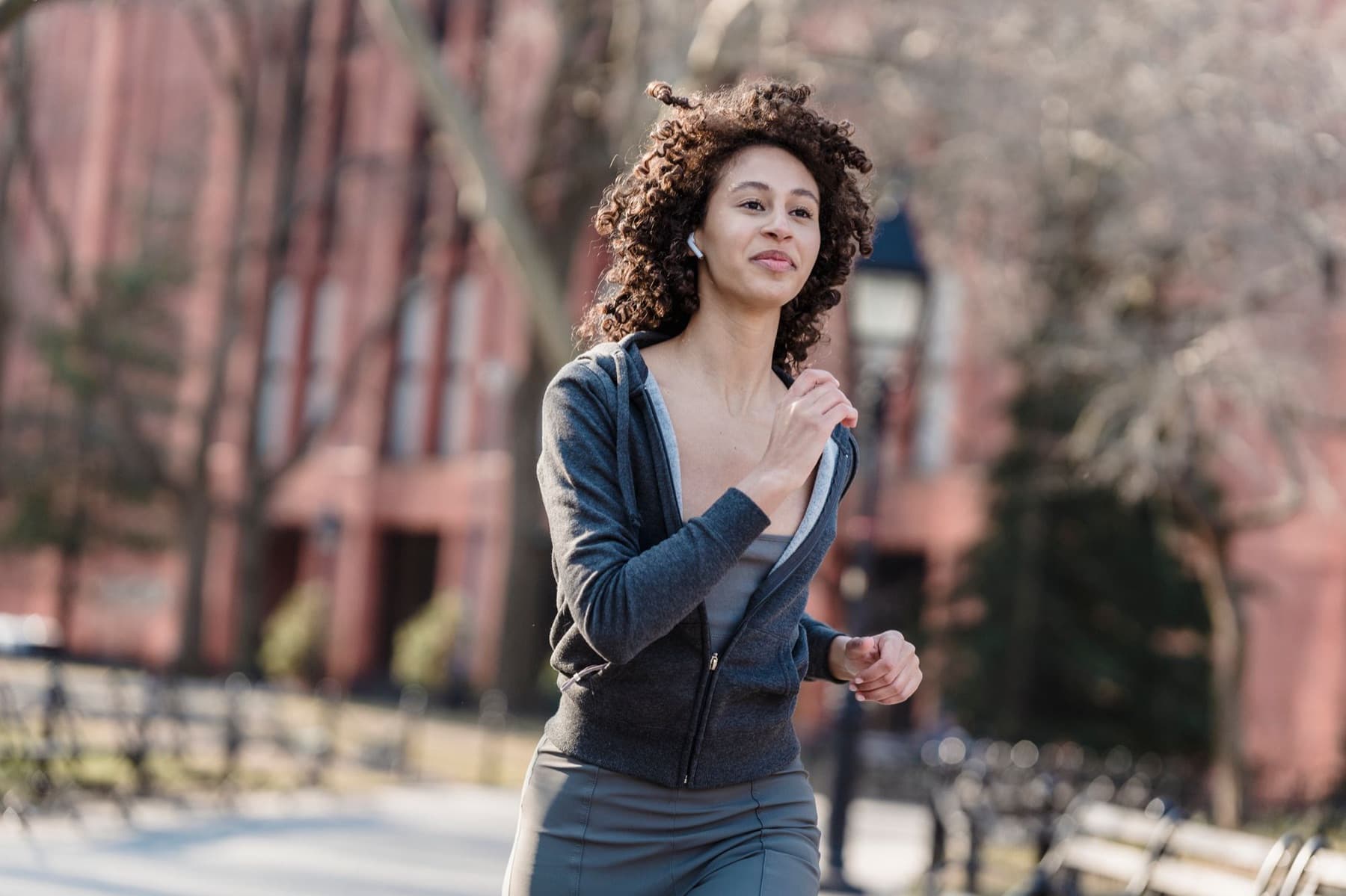 A woman with curly hair jogs through a city park in a grey zip top, wearing wireless earbuds