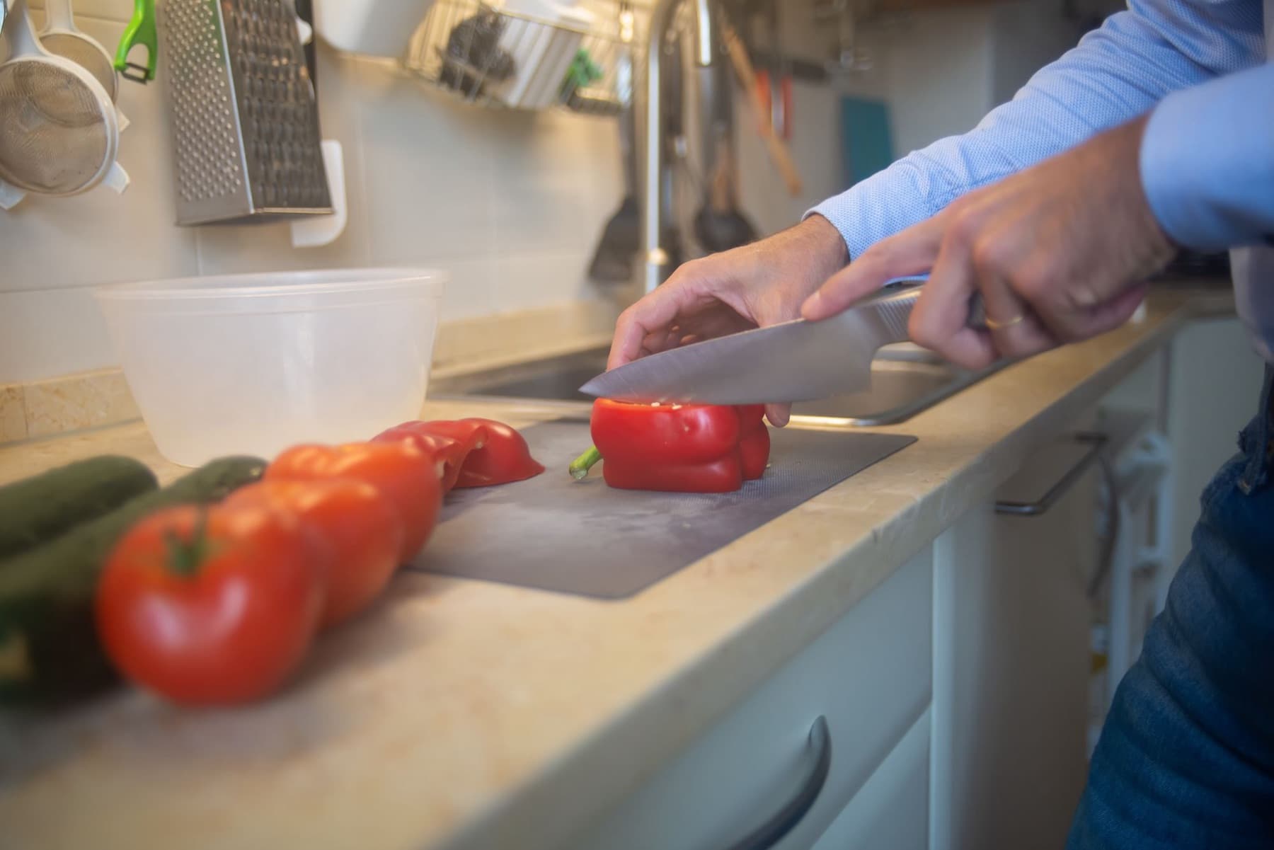 A man slices a red pepper on a chopping board at a kitchen worktop, with tomatoes and a cucumber beside him