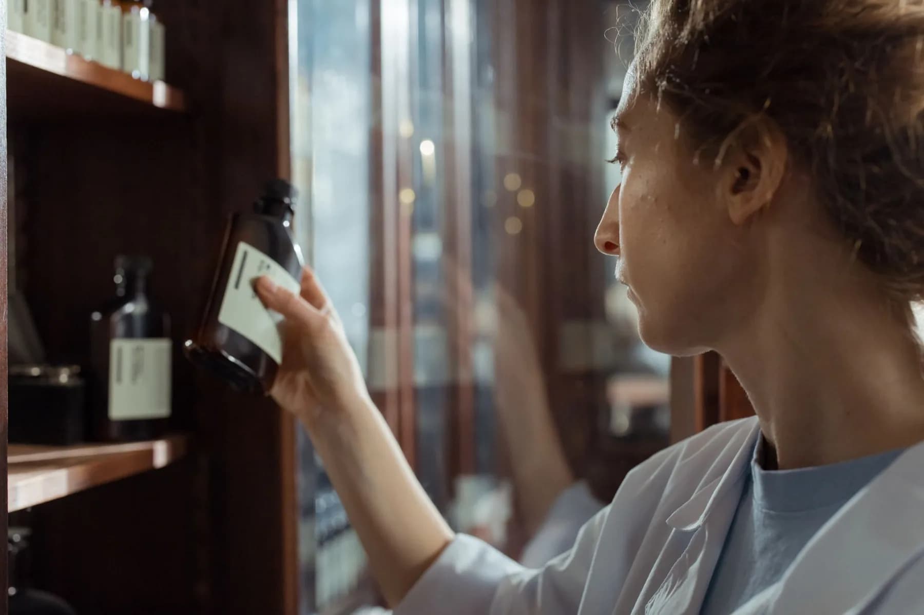 A pharmacist in a white coat reads the plain label on a brown glass bottle taken from a wooden shelf