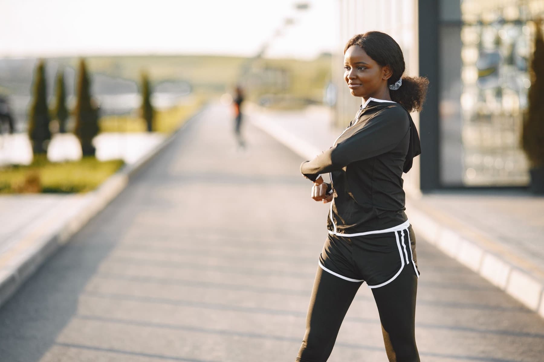 A woman in black sportswear stretches her shoulders on a paved path in low sunlight