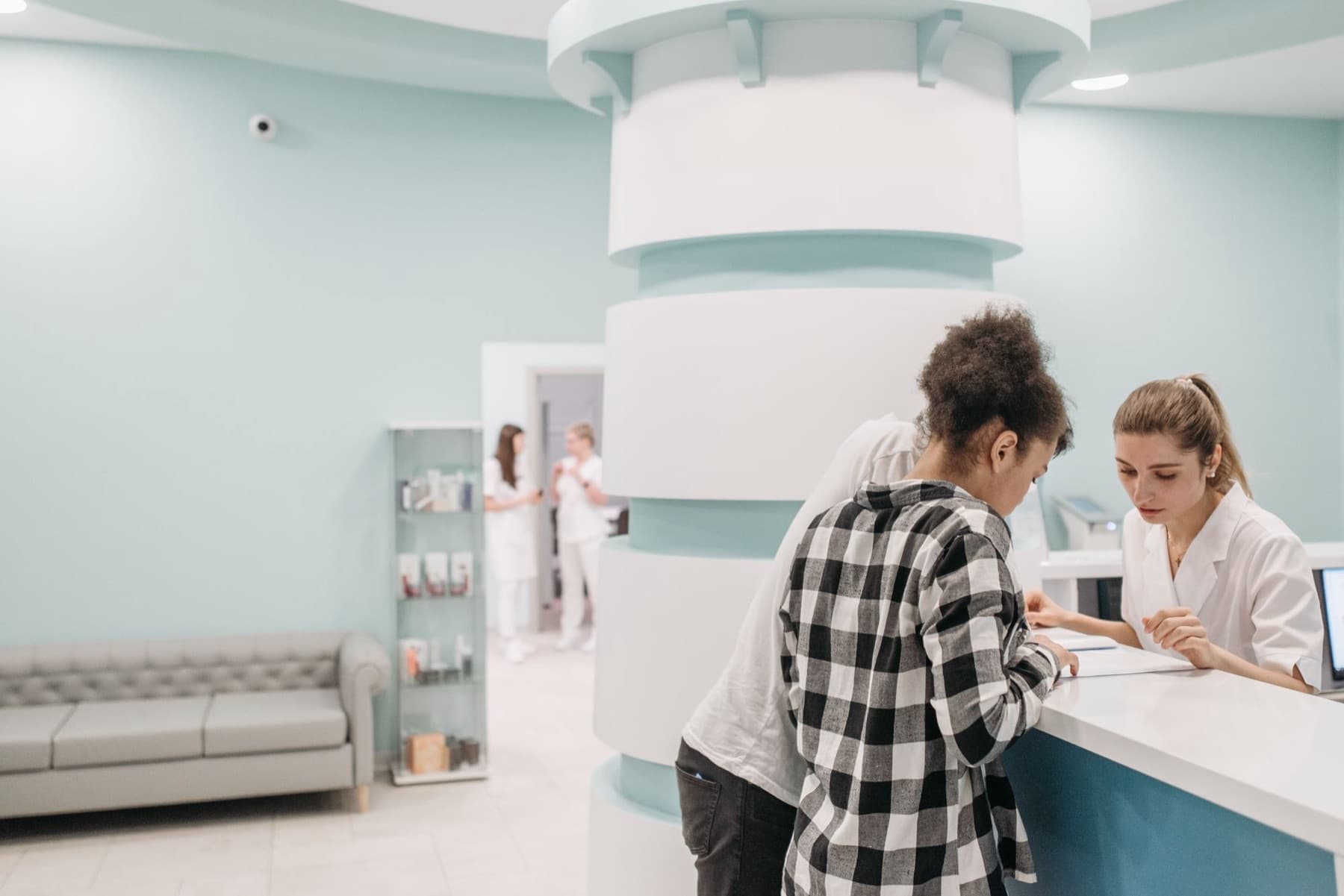 Two staff in white uniforms help a customer with paperwork at a clinic reception counter